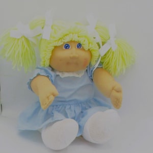 Könnte beinhalten: Eine Vintage-Puppe mit gelben Zopf-Zöpfen, die mit weißen Bändern gebunden sind. Die Puppe hat blaue Augen, ein pfirsichfarbenes Gesicht und trägt ein hellblaues Kleid mit weißen Socken. Die Puppe ist ein klassisches Spielzeug.