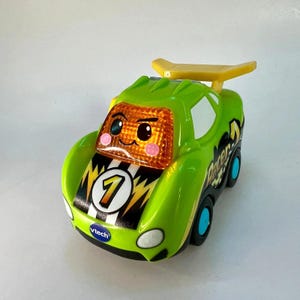 May include: A green toy race car with a yellow spoiler. The car has the number 7 on the front, a black and white checkered design, and the word "Racer" on the side. The car has blue wheels and a smiling face on the windshield.