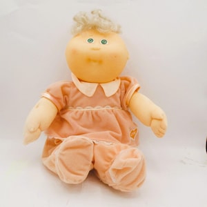 Könnte beinhalten: Eine Vintage-Puppe mit pfirsichfarbenem Gesicht, Armen und Beinen sowie hellblondem Haar. Die Puppe trägt ein pfirsichfarbenes Kleid mit weißem Kragen und Spitzensaum sowie eine passende Hose. Ein herzförmiger Aufnäher ist auf das Kleid genäht.