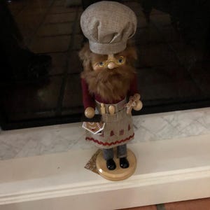 May include: A wooden nutcracker figurine dressed as a baker, wearing a brown and white chef's hat, a red apron with a white embroidered design, and holding a gingerbread cookie.