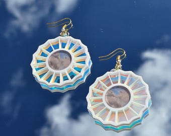 May include: A pair of iridescent white earrings with a circular design featuring a smaller circle in the center. The earrings have a gold hook.