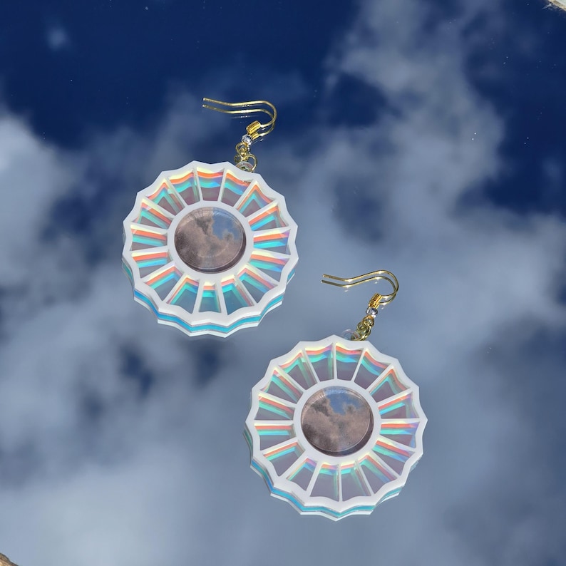May include: A pair of iridescent white earrings with a circular design featuring a smaller circle in the center. The earrings have a gold hook.