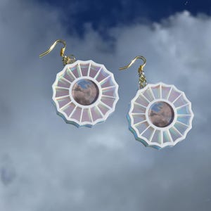 May include: A pair of iridescent white earrings with a circular design featuring a cloudy sky image in the center. The earrings have gold hooks.