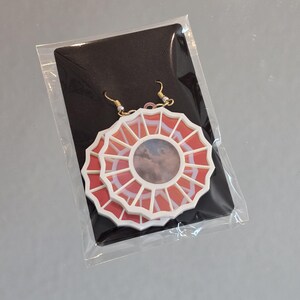 May include: A pair of white and red earrings with a circular design. The earrings feature a white frame with a red center and a cloudy image inside. The earrings are hanging from gold hooks.