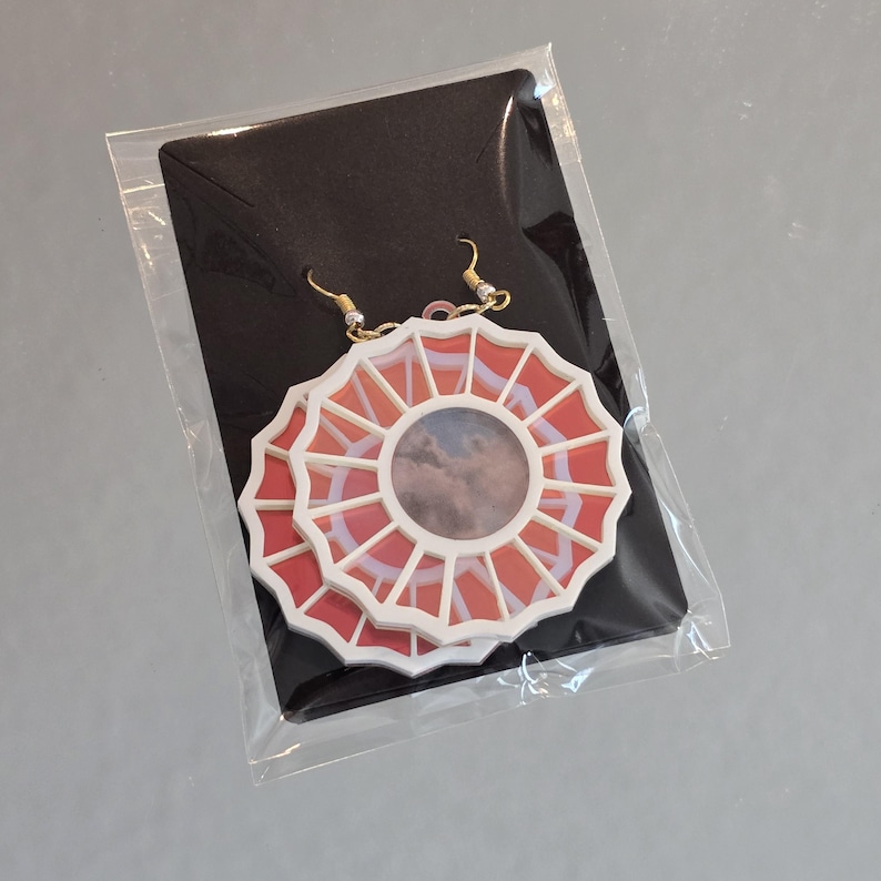 May include: A pair of white and red earrings with a circular design. The earrings feature a white frame with a red center and a cloudy image inside. The earrings are hanging from gold hooks.