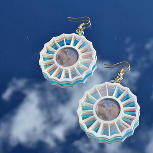 May include: A pair of iridescent white earrings with a circular design featuring a layered, multi-colored, sunburst pattern. The earrings have a gold hook closure.