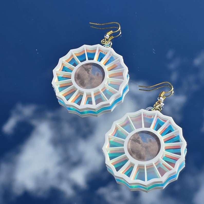 May include: A pair of iridescent white earrings with a circular design featuring a layered, multi-colored, sunburst pattern. The earrings have a gold hook closure.