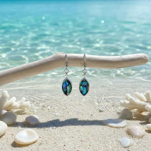 May include: A pair of silver dangle earrings with iridescent abalone shell inlays. The earrings have a teardrop shape and are suspended from a delicate chain. The earrings are displayed on a piece of driftwood against a backdrop of a sandy beach and ocean.