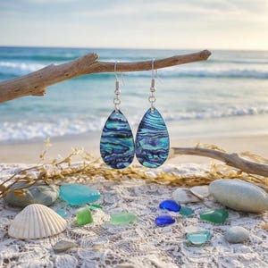 May include: A pair of teardrop-shaped earrings featuring iridescent abalone shell in shades of deep blue, teal, and purple. The earrings are suspended from silver-toned metal hooks, showcasing a natural, swirling pattern with a polished finish.