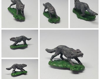 Può includere: Una statuetta di lupo in miniatura dipinta di grigio, in piedi su una base verde. Il lupo è in una posa di corsa con la bocca aperta e i denti visibili. La statuetta è alta circa 5 cm.