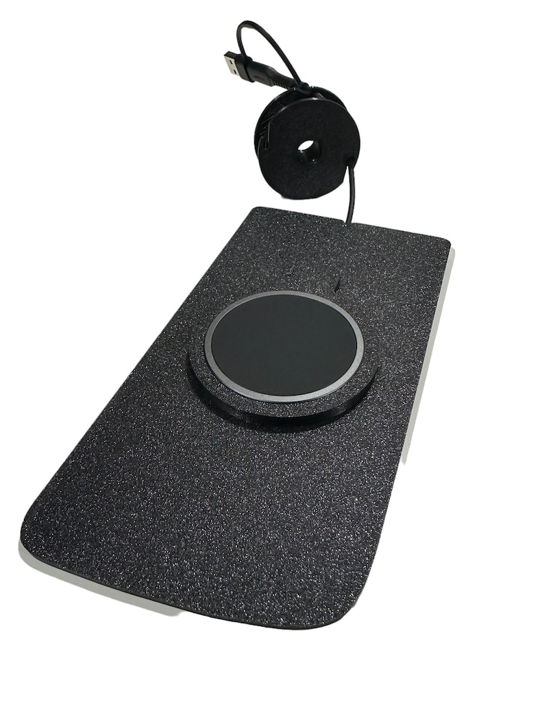 May include: A black wireless charging pad with a textured surface and a circular charging area. A black USB cable is attached and coiled. The pad is rectangular with rounded corners.