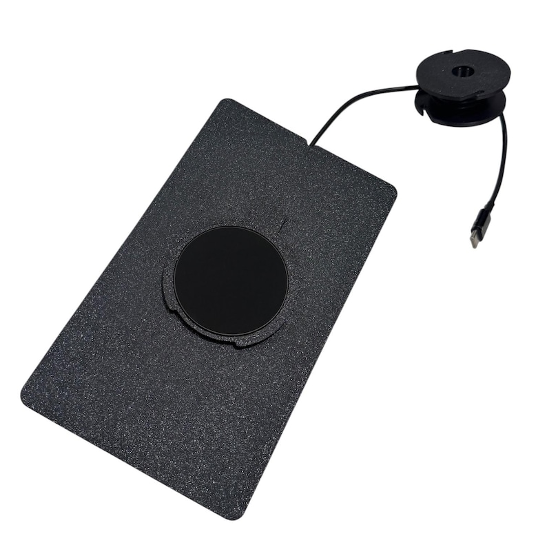 Puede incluir: Una base de carga rectangular negra con textura y una hendidura circular en el centro. Un cable negro fino se extiende desde el borde superior de la base, conect&aacute;ndose a un peque&ntilde;o componente de bobina circular negra con un conector USB-C.