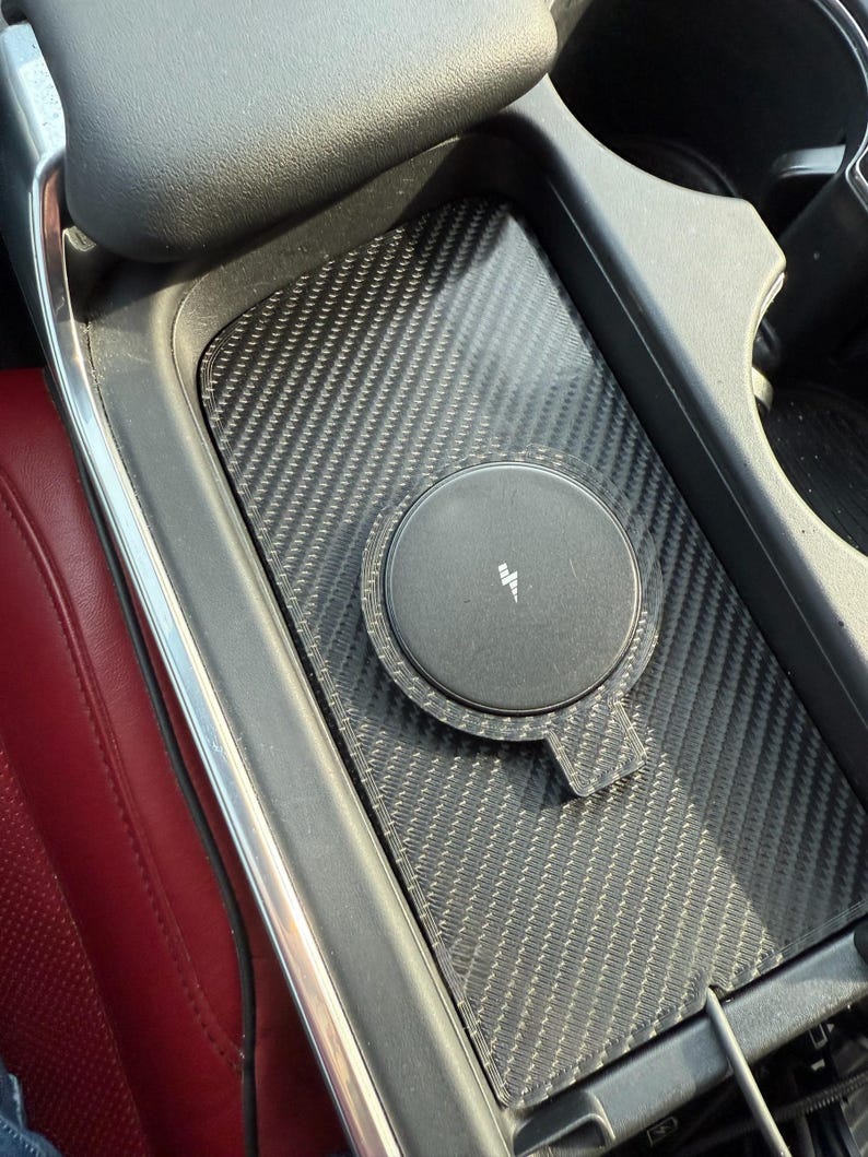 May include: A black wireless phone charger with a carbon fiber pattern is installed in a car's center console. The charger has a circular charging pad with a white lightning bolt symbol. The console is black with a red leather seat visible.