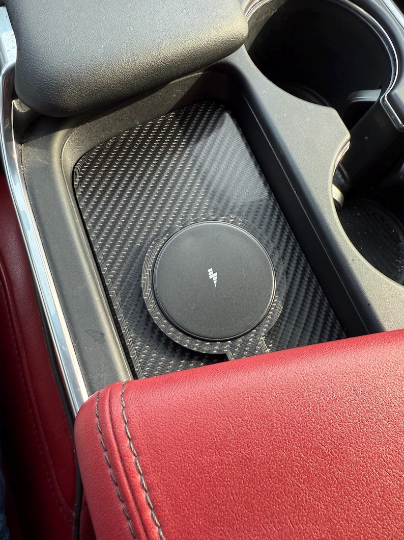 May include: A black wireless phone charger with a lightning bolt symbol sits in a car's center console. The charger is on a carbon fiber textured surface, next to a red leather armrest and cup holders.