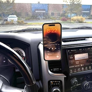 May include: A smartphone displaying 8:22 is mounted on a car dashboard. The phone, in a black case, is positioned near the air vent. The car's interior features a steering wheel, dashboard controls, and a digital display showing various settings.