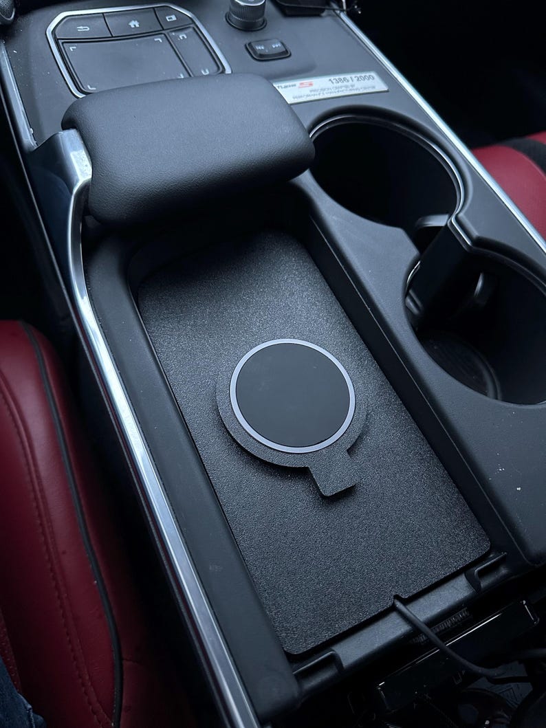 May include: A black wireless phone charger with a white outline sits in a car's center console. The charger is round with a small tab. The console is black with a textured surface, and the car's interior features red leather seats.
