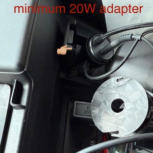 May include: Image shows a car's interior with a 12V outlet and charging cable. Text in red states, "For fast charging use the 12V outlet with minimum 20W adapter." A finger points to the outlet. A round, silver object is also visible.