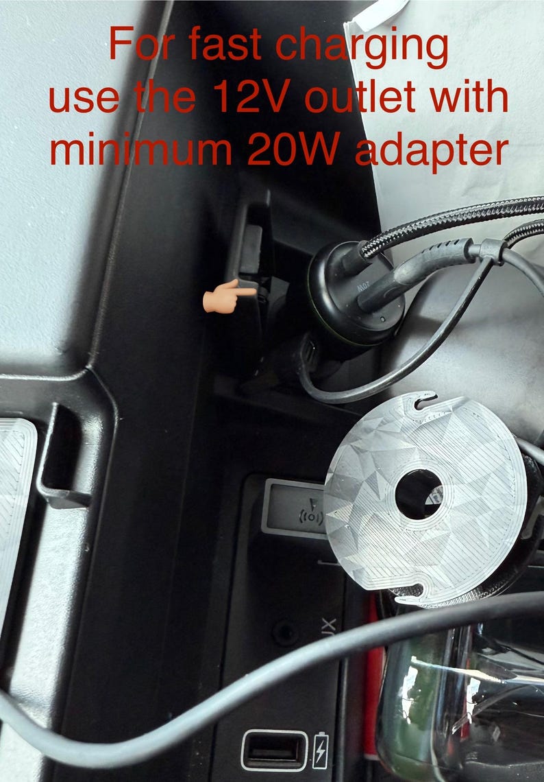May include: Image shows a car's interior with a 12V outlet and charging cable. Text in red states, "For fast charging use the 12V outlet with minimum 20W adapter." A finger points to the outlet. A round, silver object is also visible.