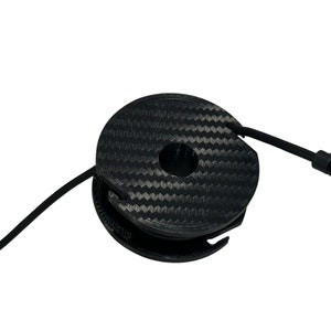May include: A black, circular cable organizer with a carbon fiber pattern. The organizer has a central hole and a slot for the cable to pass through. A black cable is partially wrapped around the organizer.