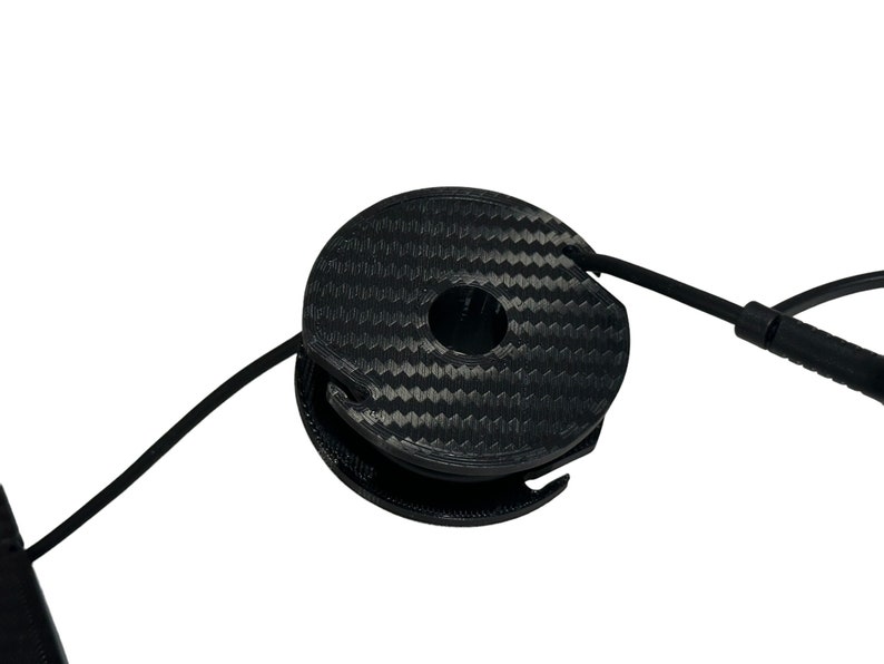 May include: A black, circular cable organizer with a carbon fiber pattern. The organizer has a central hole and a slot for the cable to pass through. A black cable is partially wrapped around the organizer.