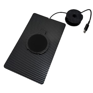 Puede incluir: Una base de carga inal&aacute;mbrica rectangular negra con textura de fibra de carbono y un centro magn&eacute;tico circular. Un cable negro fino se extiende desde el borde superior hasta una bobina circular con un conector USB-C de dise&ntilde;o oscuro.