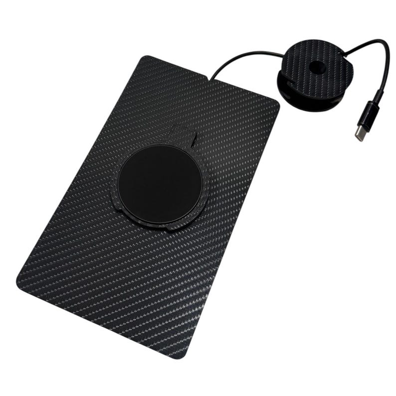 Puede incluir: Una base de carga inal&aacute;mbrica rectangular negra con textura de fibra de carbono y un centro magn&eacute;tico circular. Un cable negro fino se extiende desde el borde superior hasta una bobina circular con un conector USB-C de dise&ntilde;o oscuro.