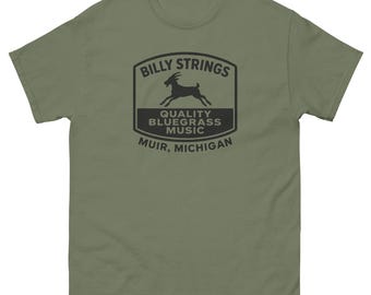May include: Olive green t-shirt featuring a black graphic with the text "Billy Strings" above a goat silhouette, and "Quality Bluegrass Music" below. The text "Muir, Michigan" is at the bottom of the design. A classic crew neck tee.
