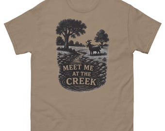 May include: A tan-colored t-shirt featuring a black and white illustration of a goat standing near a creek. The graphic includes trees, rocks, and the text "MEET ME AT THE CREEK" in a circular design.