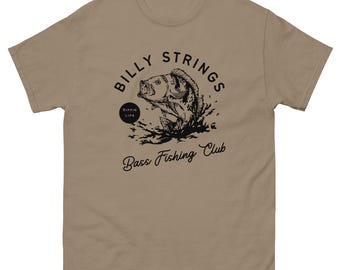 May include: A tan-colored t-shirt featuring a black graphic of a fish leaping out of water. The text "BILLY STRINGS" arches above the fish, with "Bass Fishing Club" below. A small circle reads "Rippin' Lips".