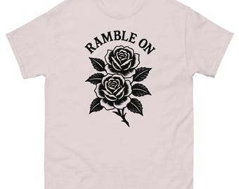 May include: A light pink t-shirt featuring a black and white floral design of two roses with leaves. The text "RAMBLE ON" is arched above the flowers. The shirt has a crew neck and short sleeves.