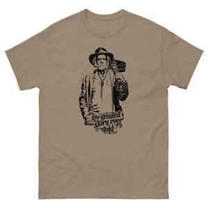 May include: A beige t-shirt with a black and white illustration of a man holding a guitar. The man is wearing a hat and jacket. The text "the greatest story ever told" is below the illustration.