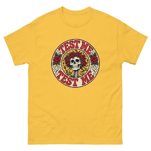 May include: A yellow t-shirt featuring a circular design with the text "TEST ME". The design includes a skull wearing a crown of red roses. The skull has a visible spine. The design is surrounded by more red roses.