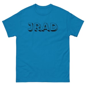 May include: A turquoise short-sleeve t-shirt with the word "JRAD" in black, 3D-style lettering across the chest. The shirt has a crew neck and a relaxed fit, suitable for casual wear.