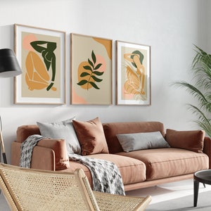 May include: Three framed abstract art prints with a minimalist style. The prints feature a green leaf, a woman in a seated pose, and a woman with her hand on her head. The prints are hung on a white wall above a brown sofa.
