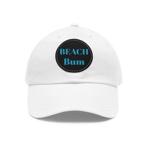 Peut inclure: Casquette de baseball blanche avec une visière incurvée. Un écusson circulaire noir sur le devant affiche les mots "BEACH Bum" en bleu clair. La casquette est en matière souple et possède une sangle réglable.