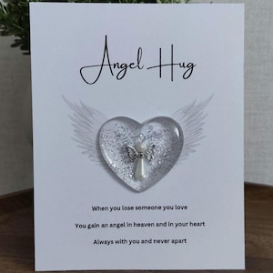 Puede incluir: Una tarjeta blanca con las palabras "Angel Hug". Un adorno transparente en forma de coraz&oacute;n con un &aacute;ngel plateado y purpurina se encuentra en la tarjeta, con alas de &aacute;ngel. La tarjeta incluye texto sobre la p&eacute;rdida y el recuerdo.