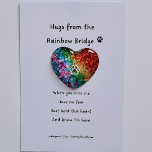 May include: A white card with the text "Hugs from the Rainbow Bridge" and a colorful heart-shaped ornament. The heart is filled with glitter and has a paw print in the center. The card also includes a poem.
