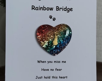 May include: A white card with the text "Hugs from the Rainbow Bridge" and a heart-shaped embellishment. The heart is filled with colorful glitter and a small silhouette of a cat. Additional text reads "When you miss me, have no fear, just hold this heart, and know I'm here."