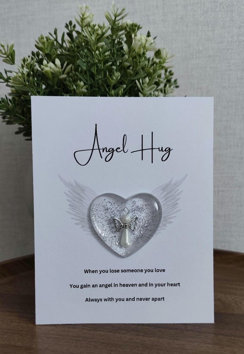 Puede incluir: Una tarjeta blanca con las palabras "Angel Hug". Un adorno transparente en forma de coraz&oacute;n con un &aacute;ngel plateado y purpurina se encuentra en la tarjeta, con alas de &aacute;ngel. La tarjeta incluye texto sobre la p&eacute;rdida y el recuerdo.