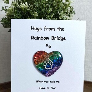 May include: A white card with the text "Hugs from the Rainbow Bridge" and a heart-shaped pendant with a paw print. The heart is filled with glitter in rainbow colors. The card also includes the text "When you miss me, Have no fear, Just hold this heart, And know I'm here."