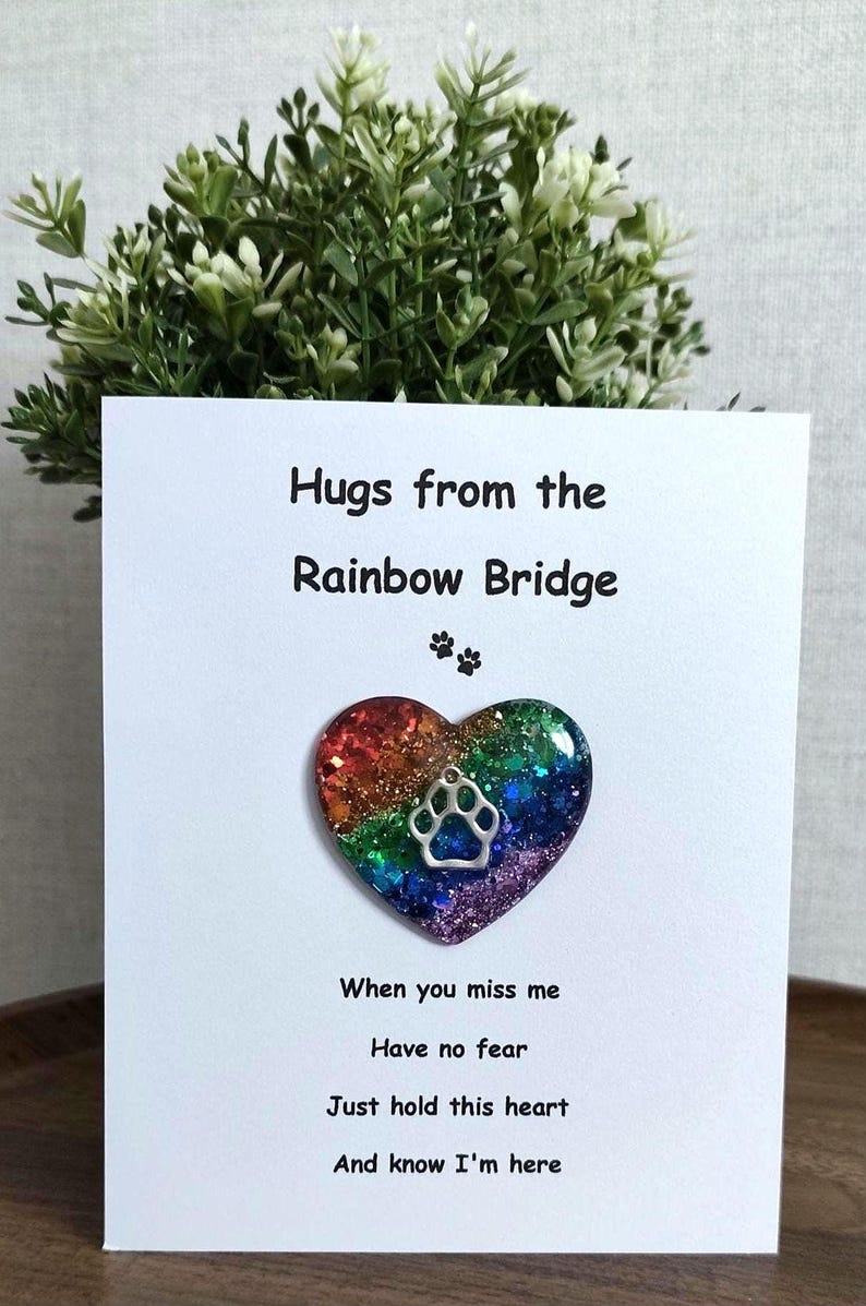 May include: A white card with the text "Hugs from the Rainbow Bridge" and a heart-shaped pendant with a paw print. The heart is filled with glitter in rainbow colors. The card also includes the text "When you miss me, Have no fear, Just hold this heart, And know I'm here."