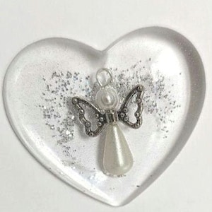 Puede incluir: Un pisapapeles de resina transparente en forma de coraz&oacute;n con purpurina plateada y un peque&ntilde;o dije de &aacute;ngel. El dije de &aacute;ngel presenta una cabeza y un cuerpo en forma de l&aacute;grima de perla, con alas plateadas ornamentadas.