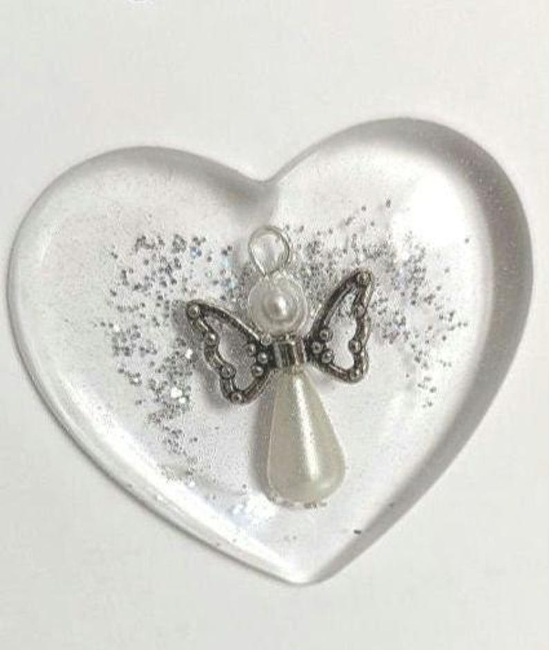 Puede incluir: Un pisapapeles de resina transparente en forma de coraz&oacute;n con purpurina plateada y un peque&ntilde;o dije de &aacute;ngel. El dije de &aacute;ngel presenta una cabeza y un cuerpo en forma de l&aacute;grima de perla, con alas plateadas ornamentadas.
