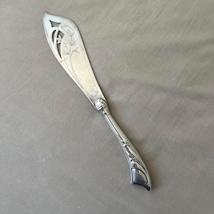 May include: A silver-toned metal serving knife featuring an elongated, pointed blade with a decorative cutout near the tip and an engraved floral motif. The handle is curved and detailed with embossed botanical elements and a smooth, polished finish.