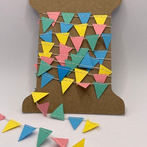 May include: A spool of colourful paper triangle bunting with a white string. The bunting is in shades of yellow, blue, green, and pink.