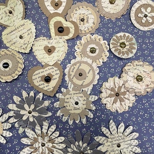 May include: Assortment of paper craft embellishments in various shapes and sizes. The embellishments are made of paper with a vintage text pattern and feature brown and gray colors. Some embellishments have a decorative button in the center.