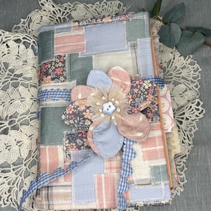 May include: A handmade journal with a patchwork cover in pastel colors, including pink, blue, and floral patterns. A large fabric flower with a button center adorns the front. Blue gingham ribbon ties the journal closed.