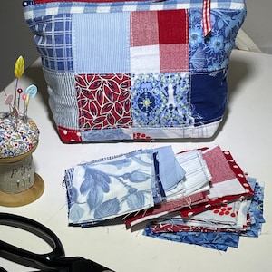 Könnte beinhalten: Ein Patchwork-Etui mit Reißverschluss in Blau-, Rot- und Weißtönen. Die Tasche weist eine Vielzahl von Stoffmustern auf, darunter Vichy, Blumenmuster und unifarbene Farben. Ein kleines Nadelkissen mit bunten Nadeln und eine schwarze Schere sind ebenfalls zu sehen.