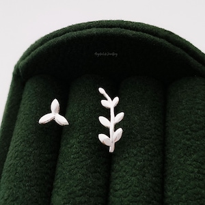 May include: A pair of silver-colored stud earrings. One earring features a three-petal design, and the other has a branch with leaves. The earrings are displayed on a dark green, textured surface.