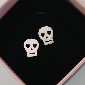 May include: A pair of silver skull-shaped stud earrings. Each earring features heart-shaped eye cutouts and a jagged mouth. The earrings are presented on a black surface within a pink box.